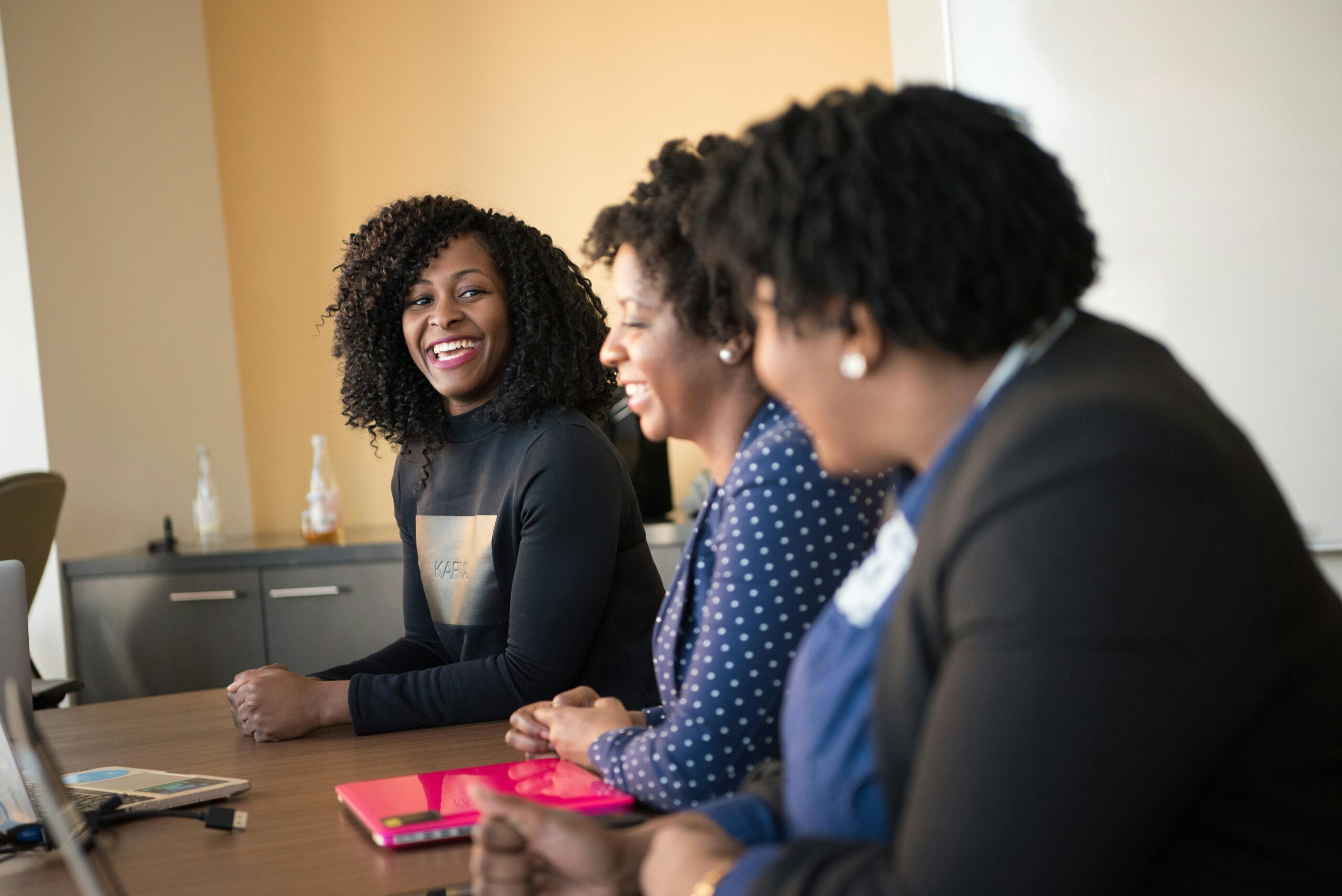 Three Black women in professional attire laughing and collaborating during a business meeting with laptops on a wooden table.