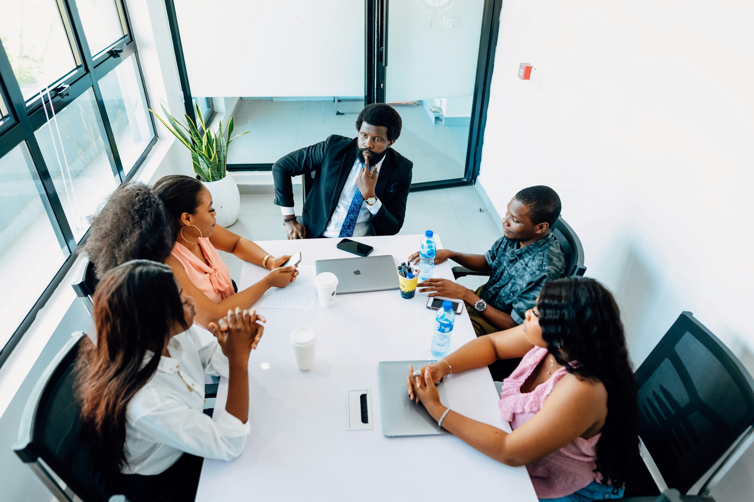 A diverse group of five Black professionals sitting around a white conference table in a bright office during a strategy meeting.